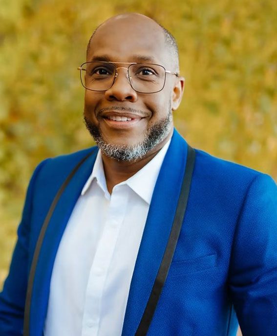 Smiling man in blue blazer and glasses against a warm, blurred background