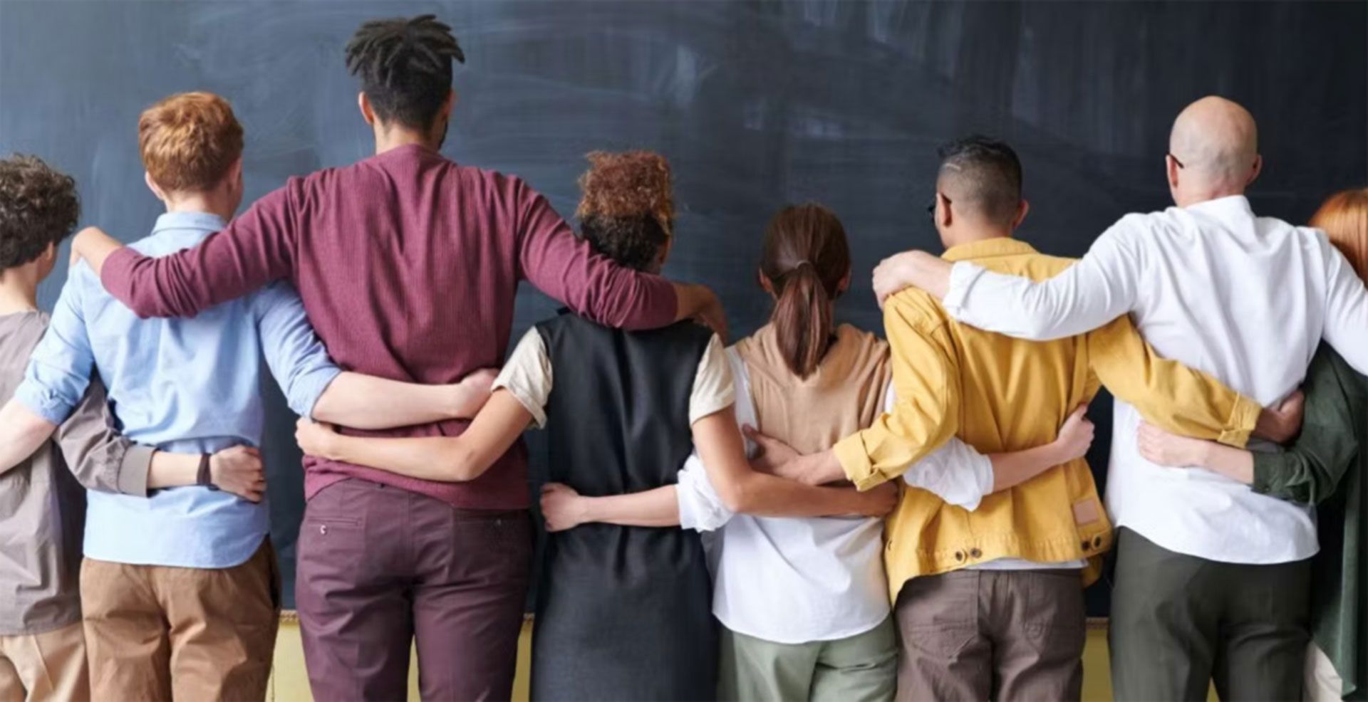 Group of people standing arm-in-arm in front of a chalkboard, seen from behind
