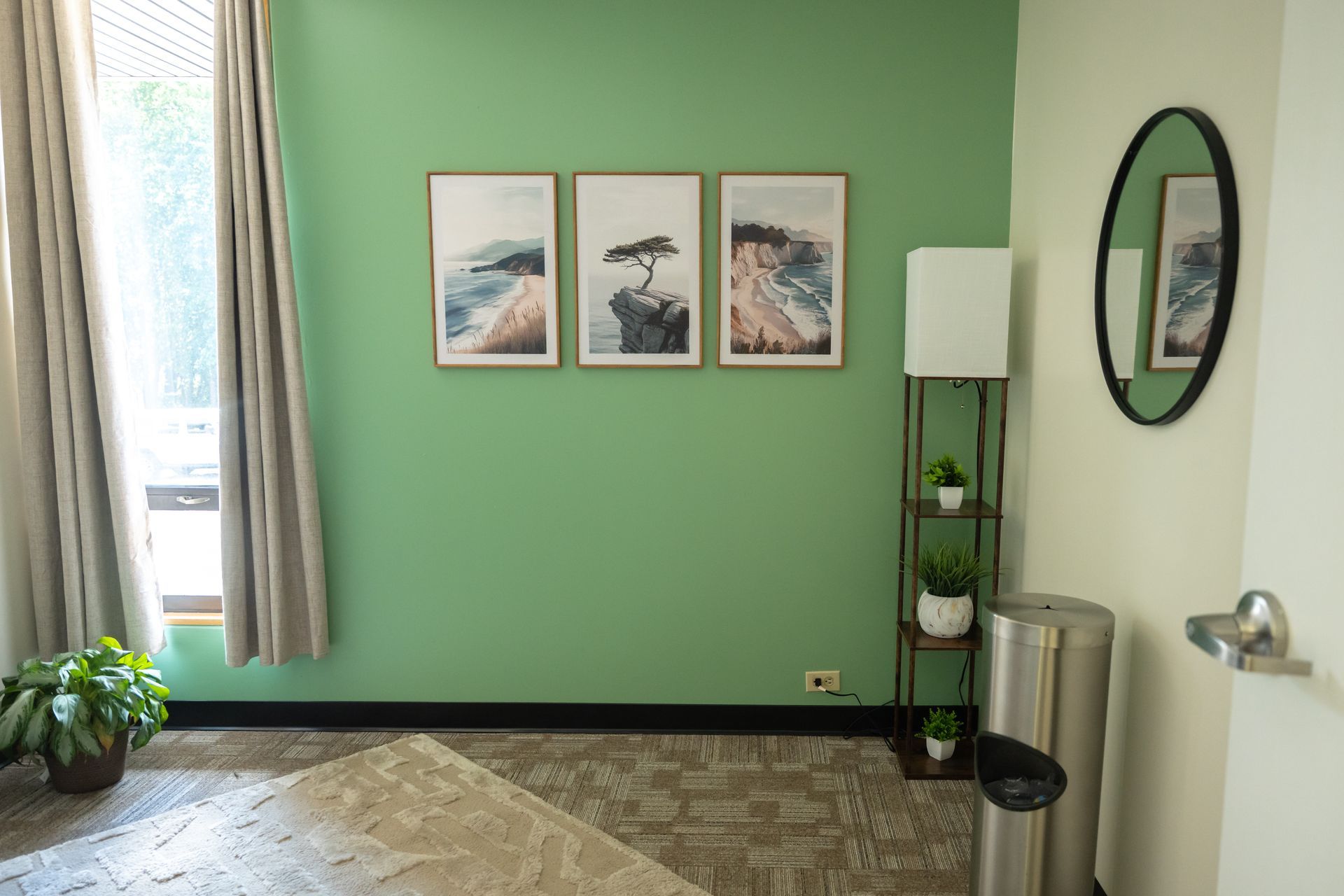 Green room with framed art, lamp, plant stand, trash can, and patterned rug near a window