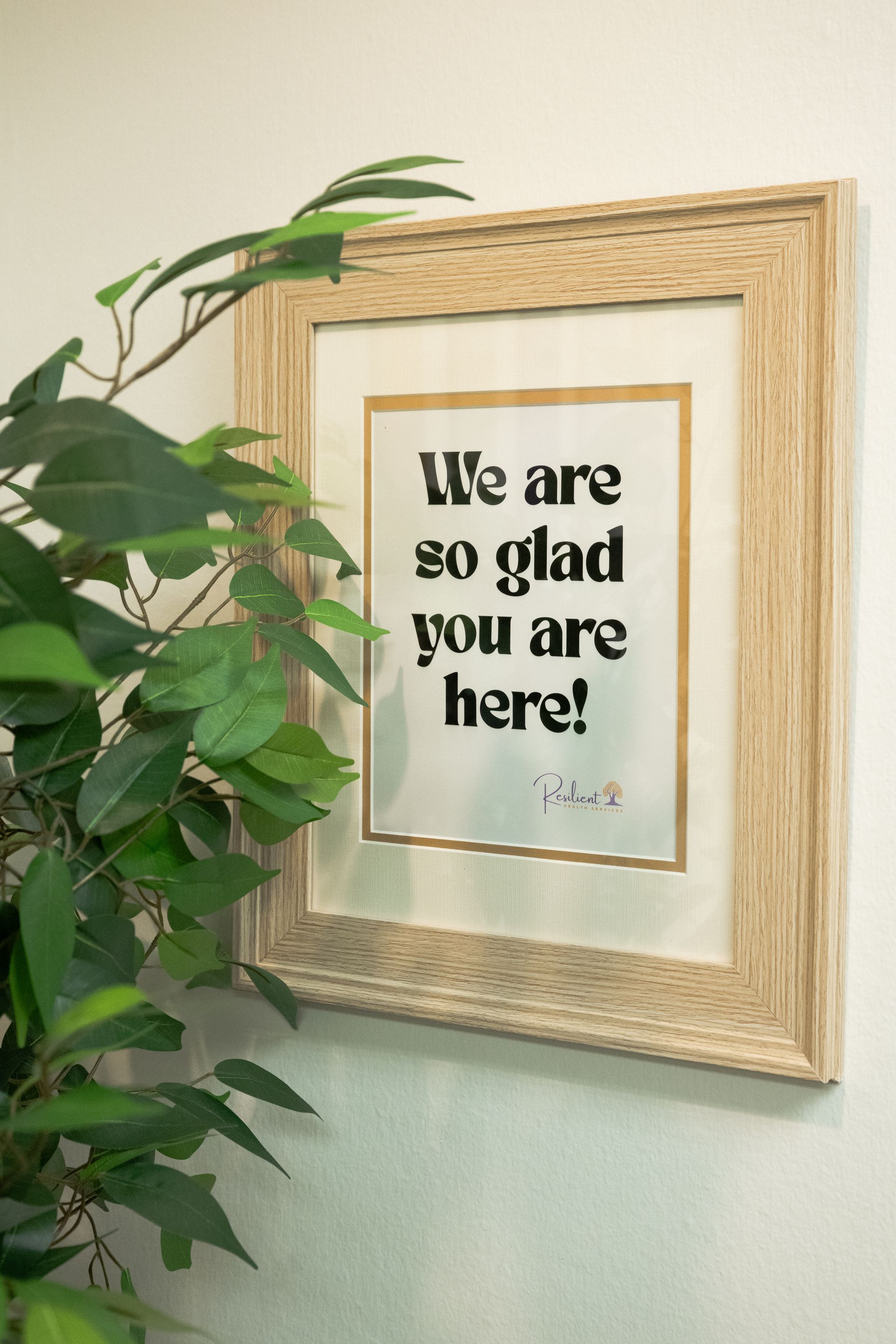 Framed “We are so glad you are here!” sign beside a leafy green plant on a wall