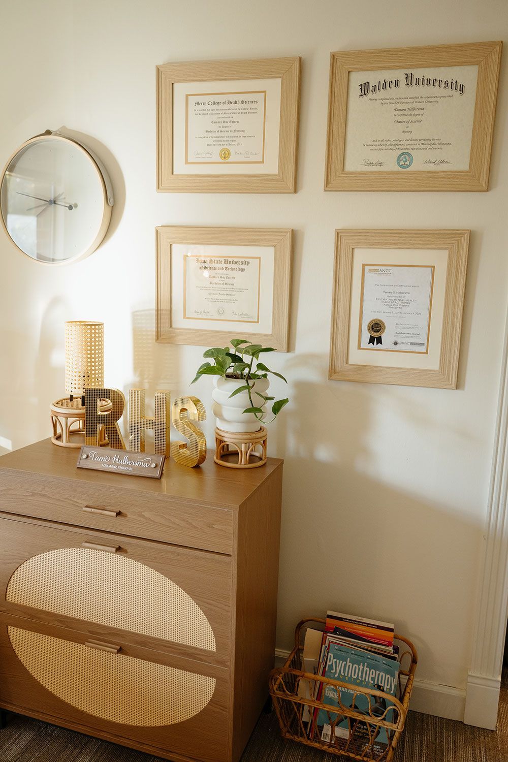 Cozy corner with framed certificates, round mirror, lamp, plant, and woven cabinet in warm beige tones