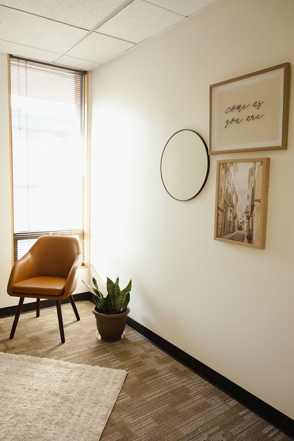 Small waiting area with an orange chair, potted plant, round mirror, and framed wall art by a window.