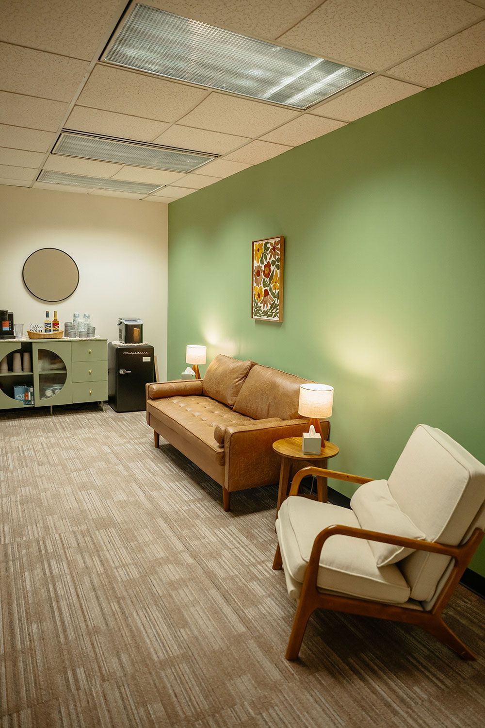 Small waiting room with tan sofa, white armchair, green wall, and patterned carpet under fluorescent lights