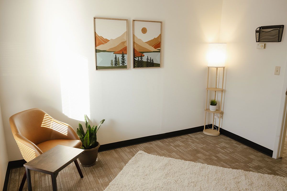Minimalist living room with wicker chair, potted plant, floor lamp, and two framed landscape prints.
