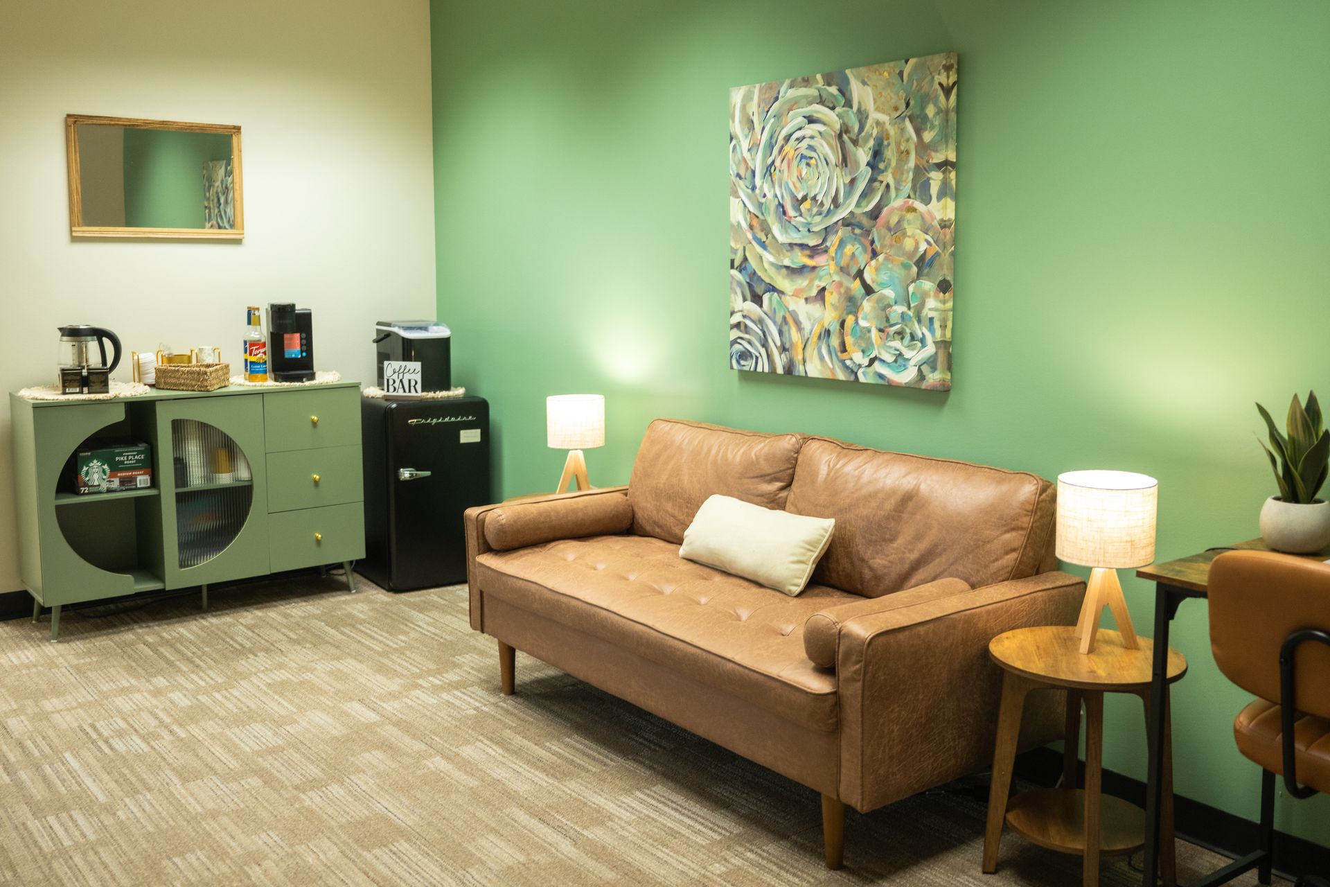 Cozy office lounge with green walls, tan sofa, side tables, lamps, and a cabinet with books and decor.