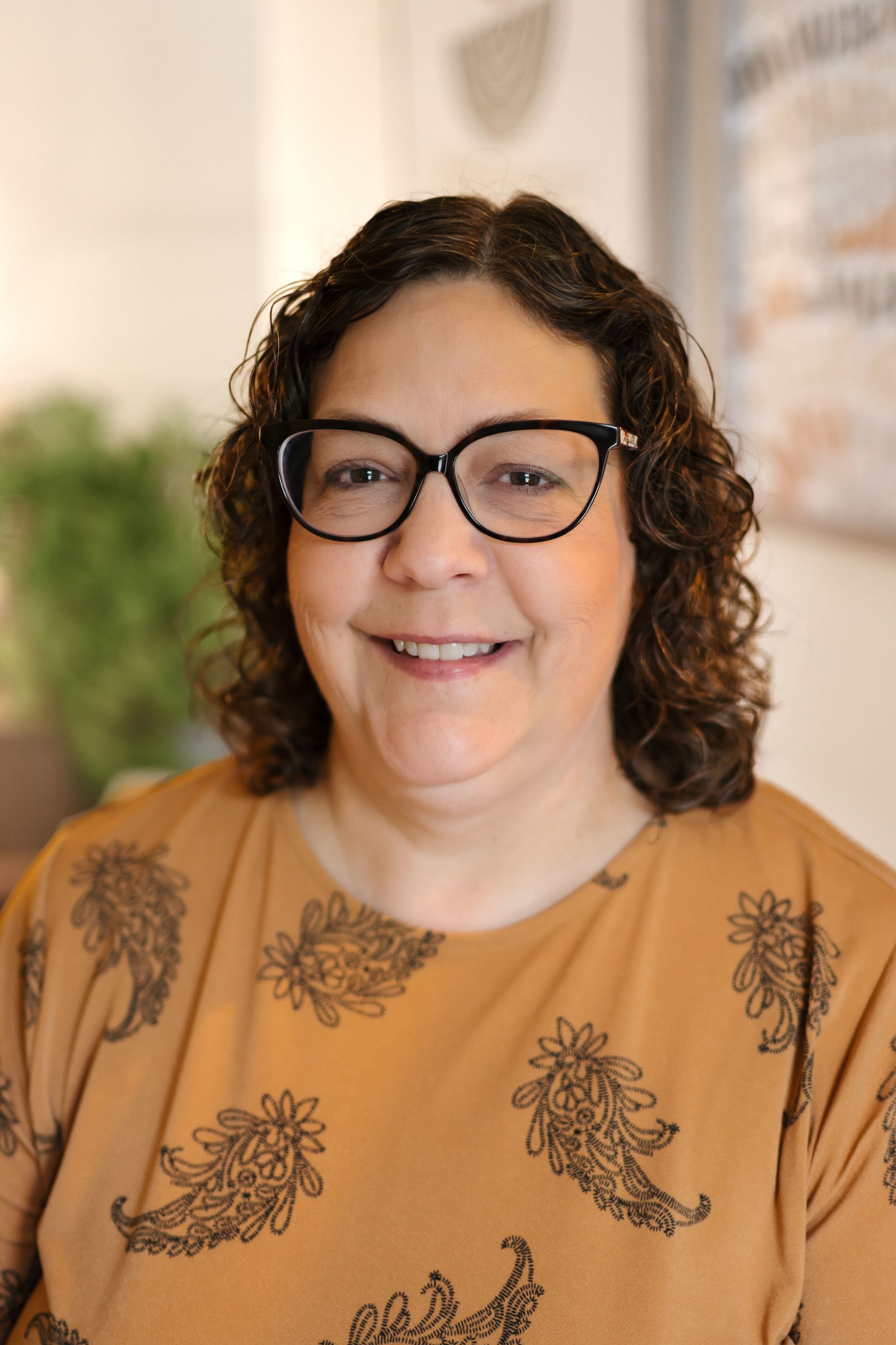 Smiling person with curly hair and glasses wearing a tan floral top against a blurred indoor background