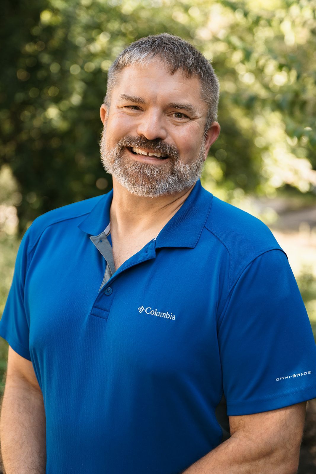 Smiling man in a blue polo shirt standing outdoors with a blurred green background