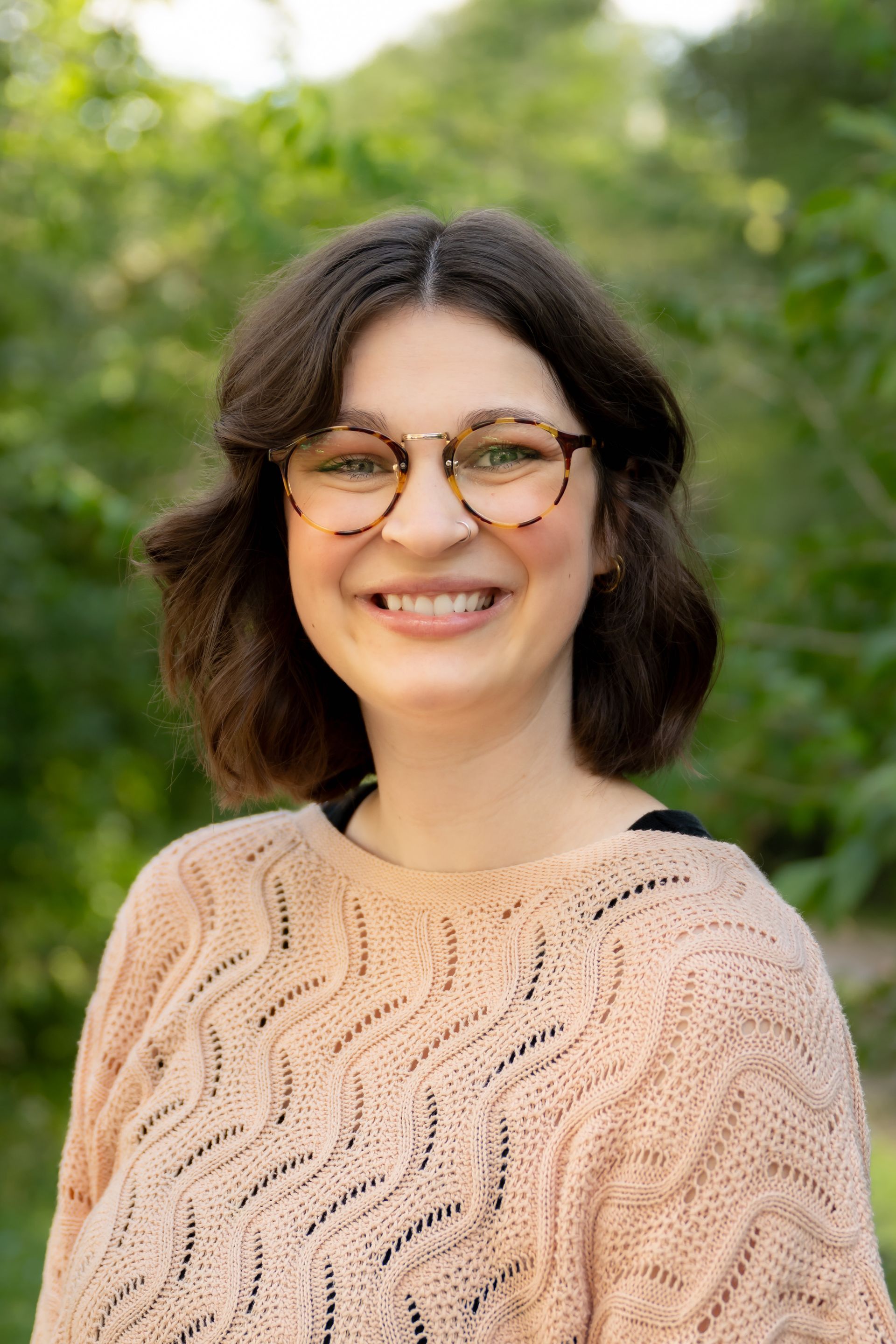 Smiling person with glasses in a peach sweater outdoors with blurred greenery background