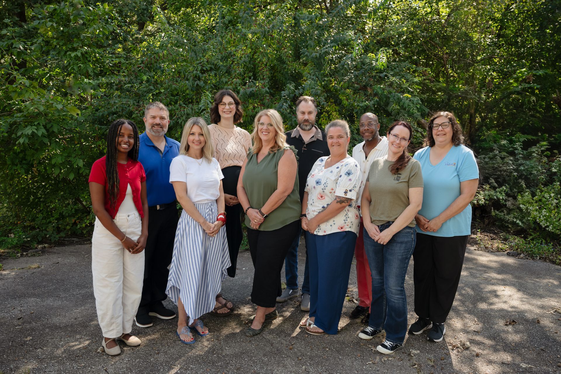 Ten people standing together outdoors in front of green trees, smiling at the camera.