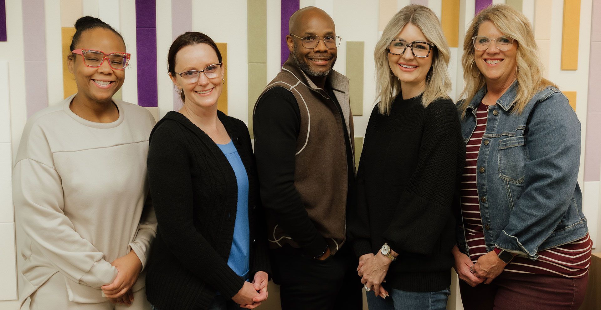 Five smiling people standing indoors, posed together in front of striped walls.