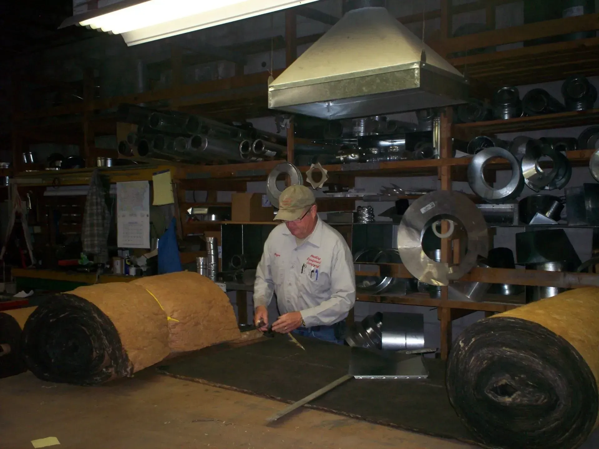 A person wearing a tan cap and work shirt works at a shop table with insulation rolls and metal ductwork parts on shelves.