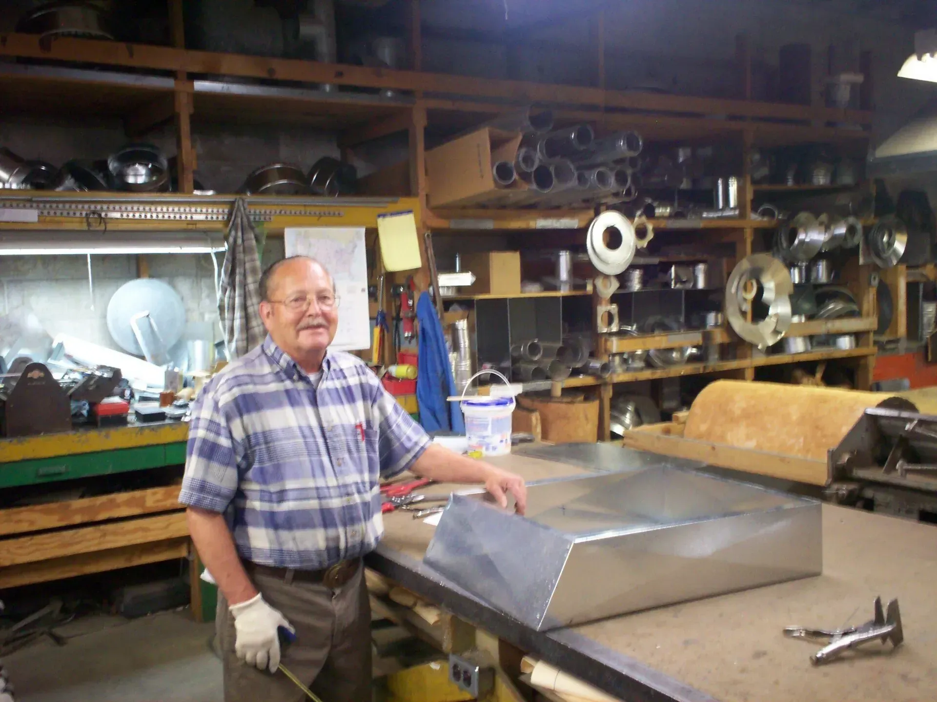A worker in a plaid shirt stands in a metal workshop, touching a sheet metal project on a workbench.