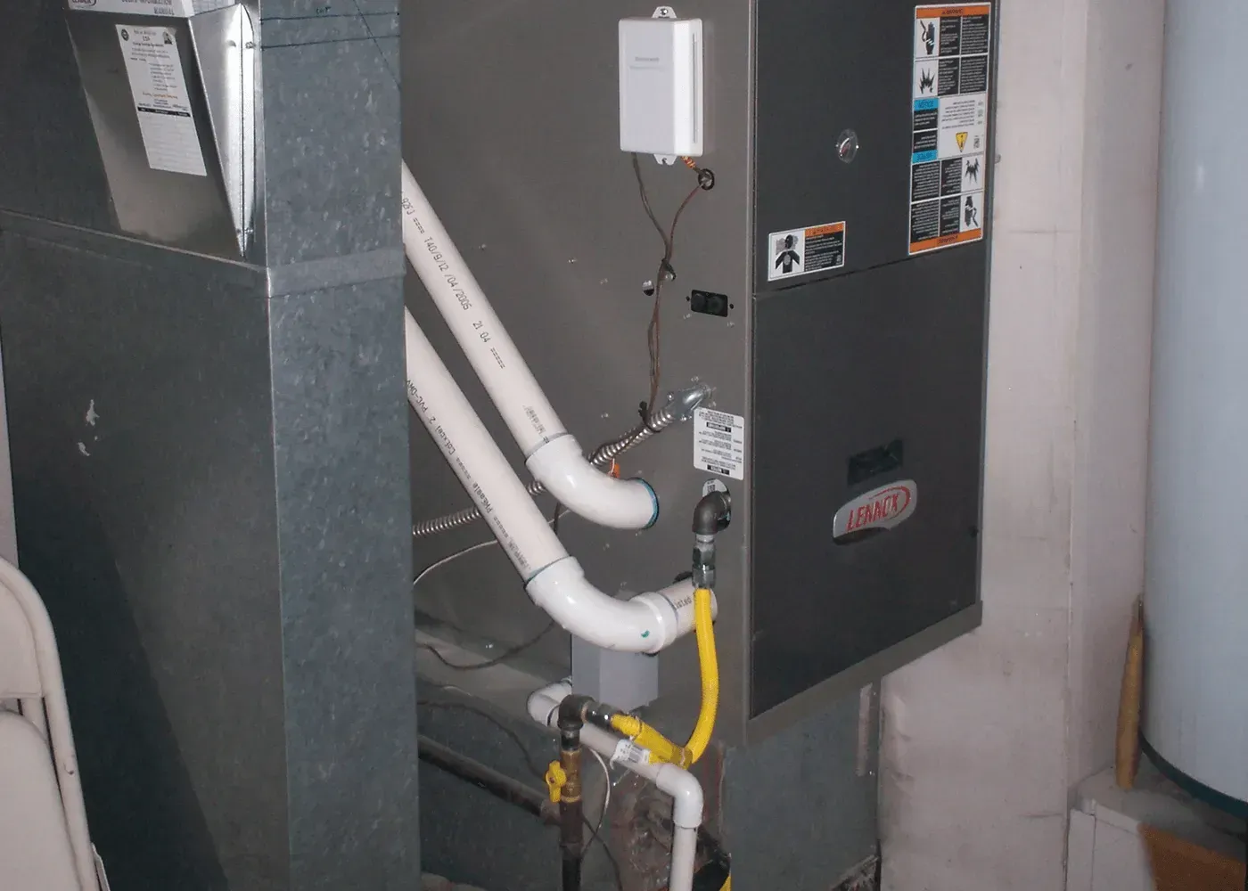 A gray Lennox furnace with two white PVC pipes and a yellow gas line connected to the side in a utility space.