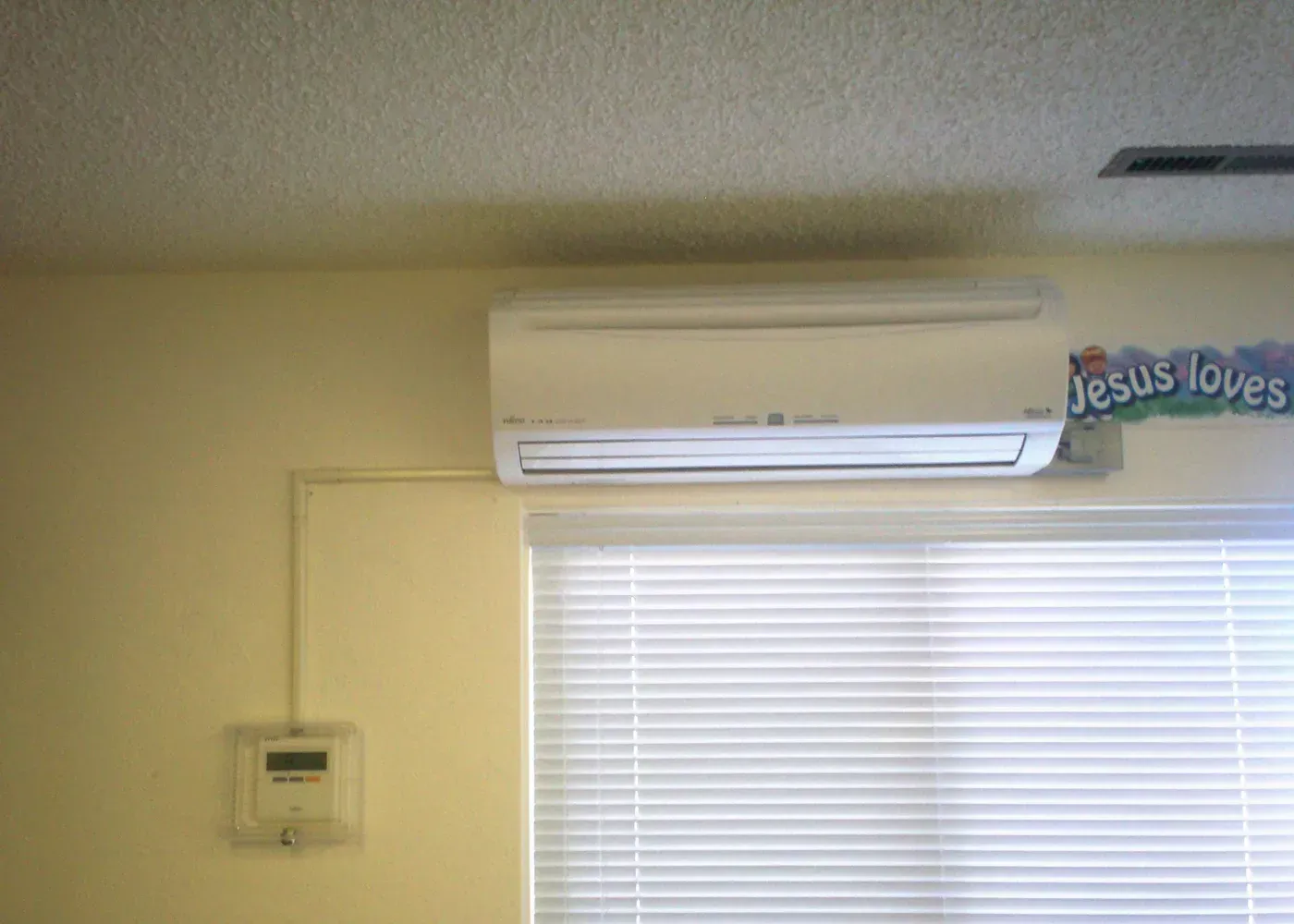 A white wall-mounted air conditioning unit is positioned above a window with closed blinds, with a thermostat below it.
