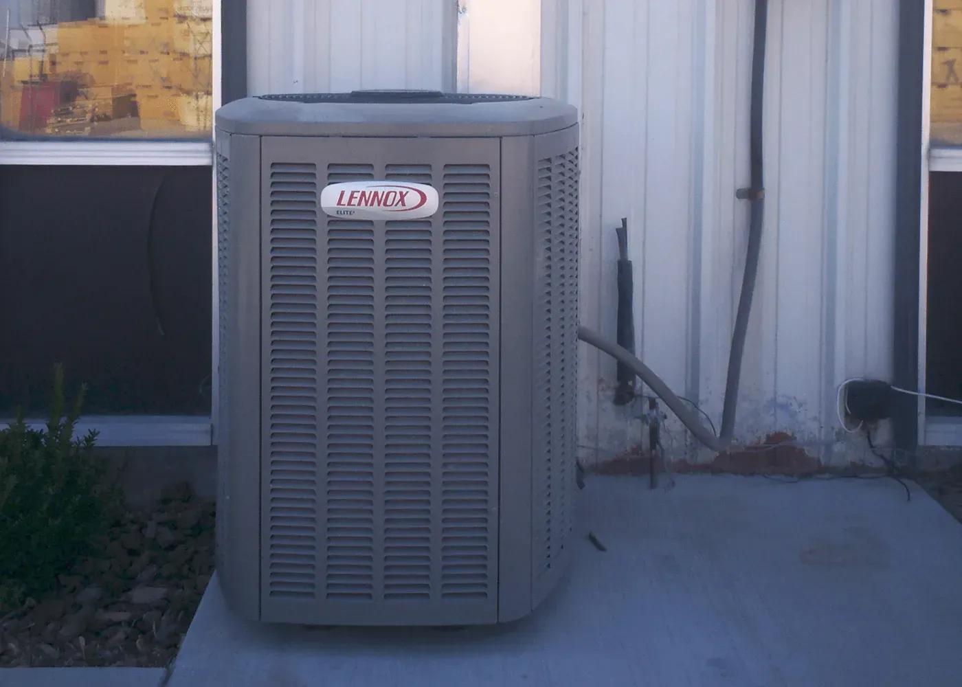 A grey Lennox outdoor HVAC unit sitting on a concrete pad against a metal building wall.