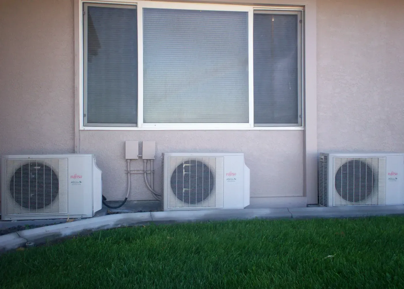 Three white air conditioning condenser units sit on the ground in front of a beige wall with a large window and green grass.