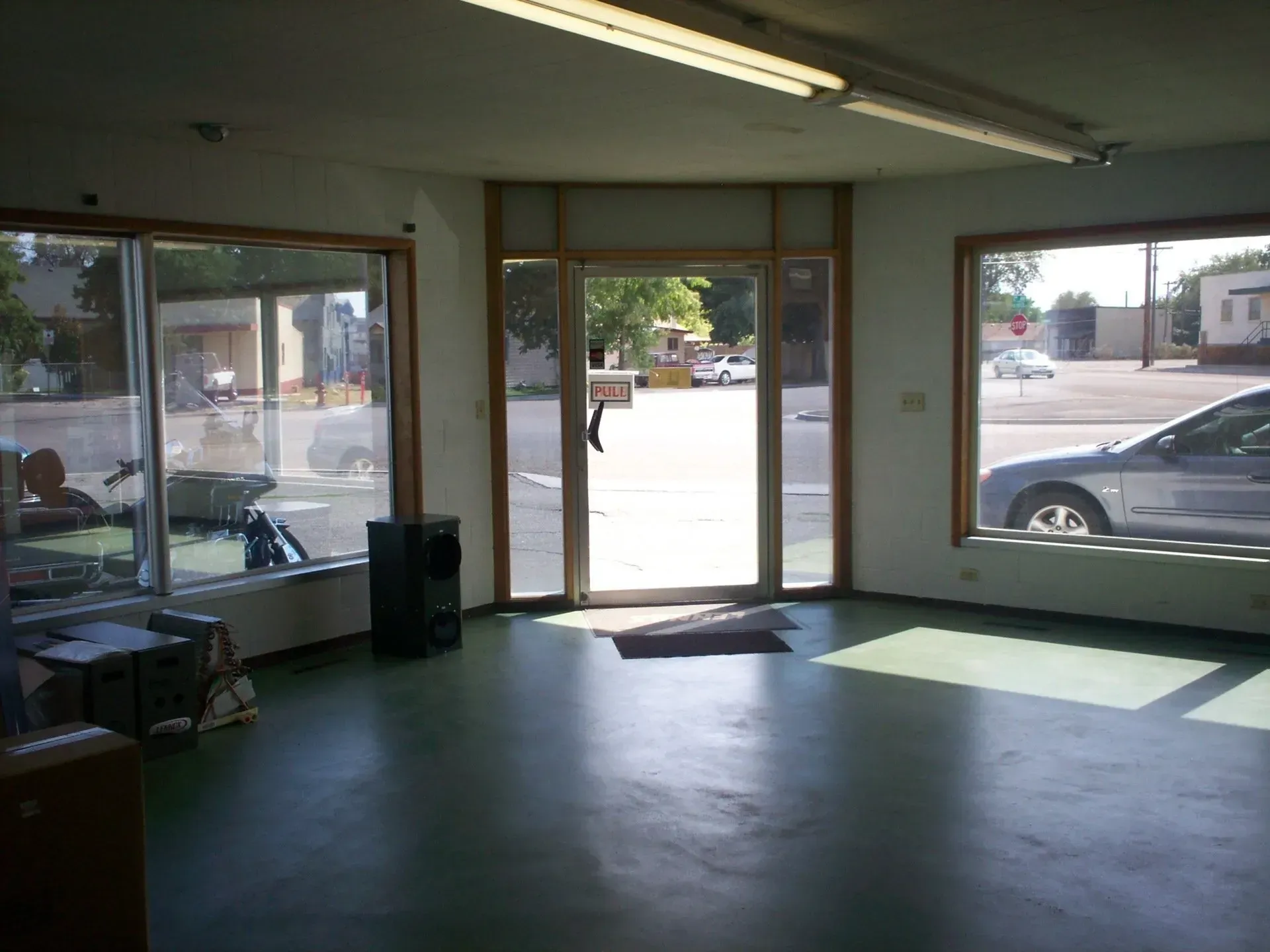 An interior view of a commercial storefront with large windows, an entrance door, green flooring, and overhead lighting.