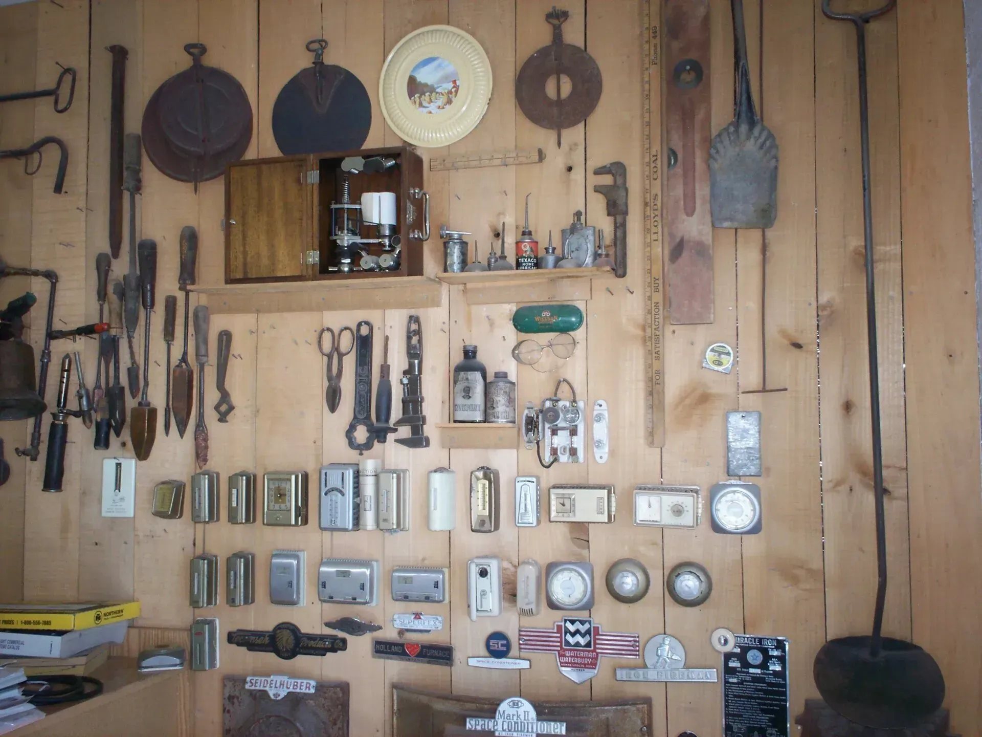 A collection of vintage tools, mechanical parts, and small metal objects mounted on a light-colored wood paneled wall.