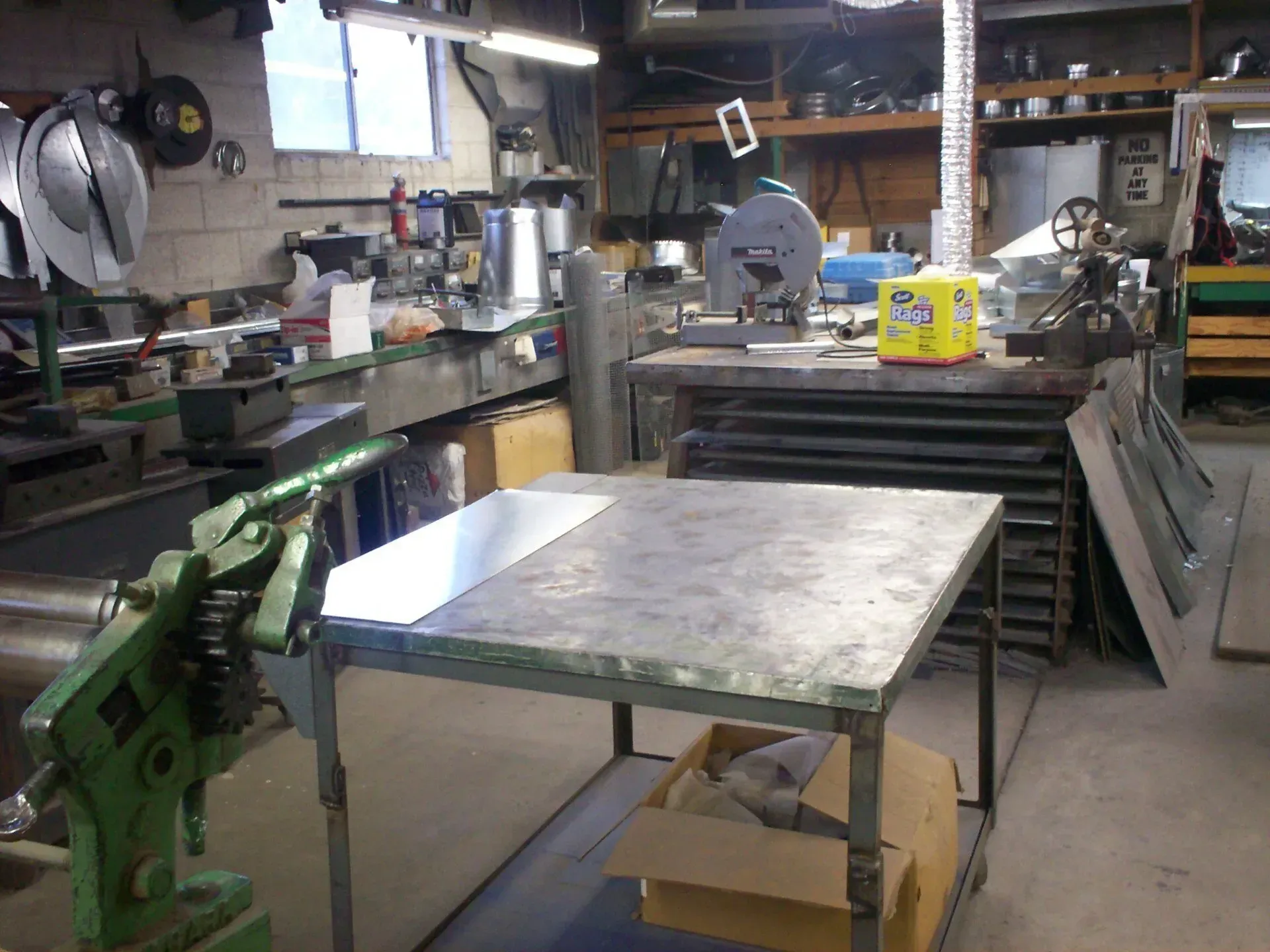 A cluttered metalworking shop features a central steel workbench, sheet metal, and specialized machinery.