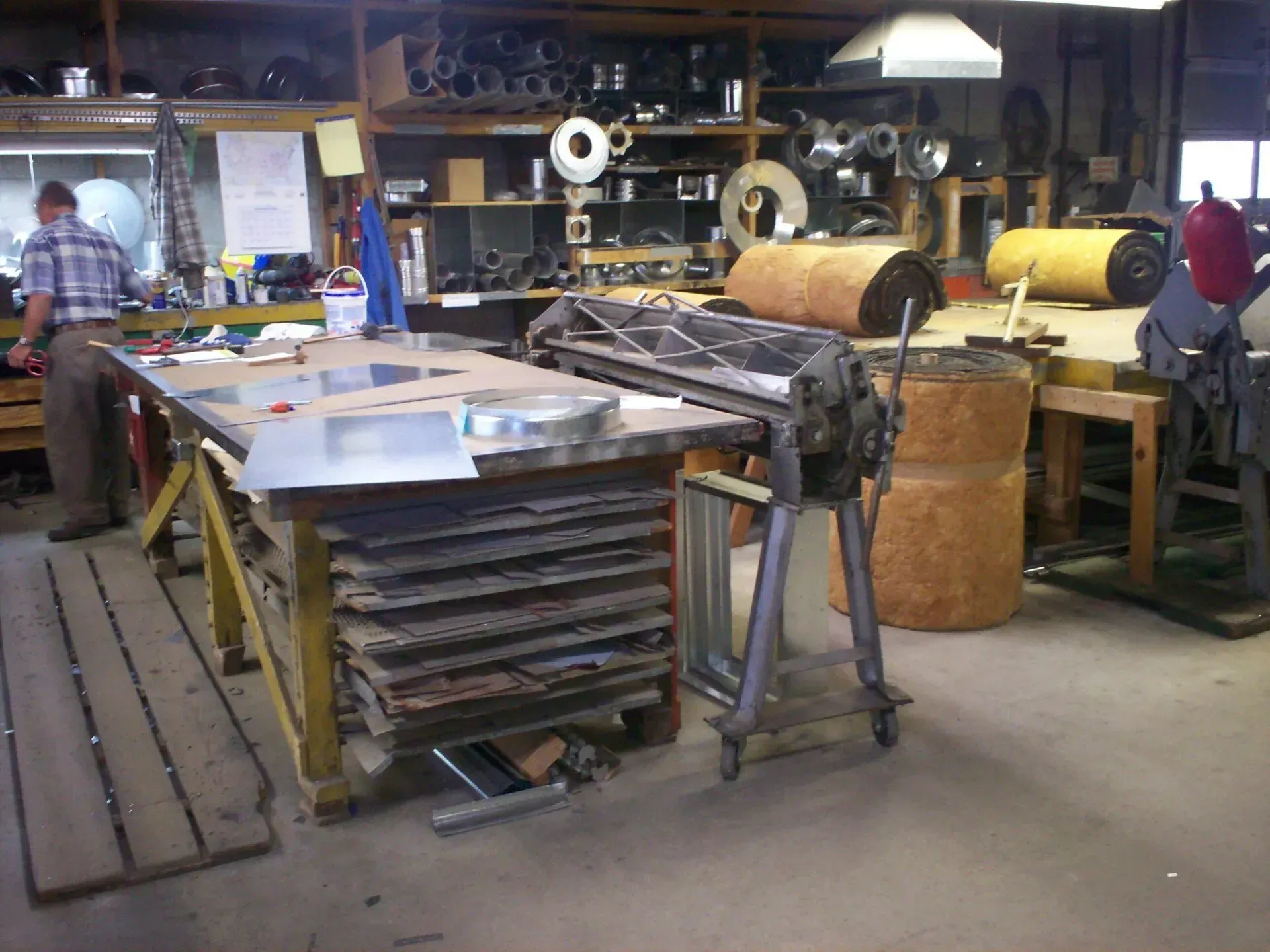 A person works at a table in a sheet metal workshop equipped with industrial tools, materials, and storage shelving.