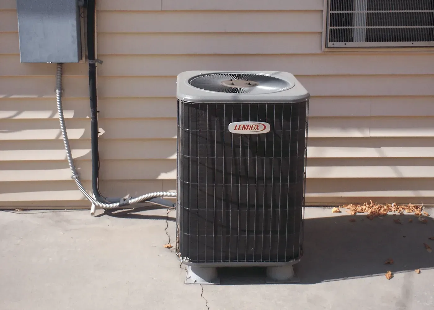A gray Lennox air conditioning unit sits on a concrete pad against the beige siding of a house.