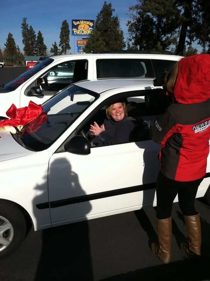 Woman in white car waving, red bow on the hood. Another person in red jacket stands next to the car.