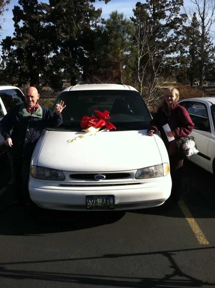 Man and woman stand beside a white car with a red bow, possibly a gift, in a parking lot.