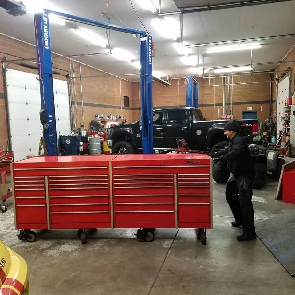 Man pushing a large red tool chest on wheels in an auto repair shop. A black truck is in the background.