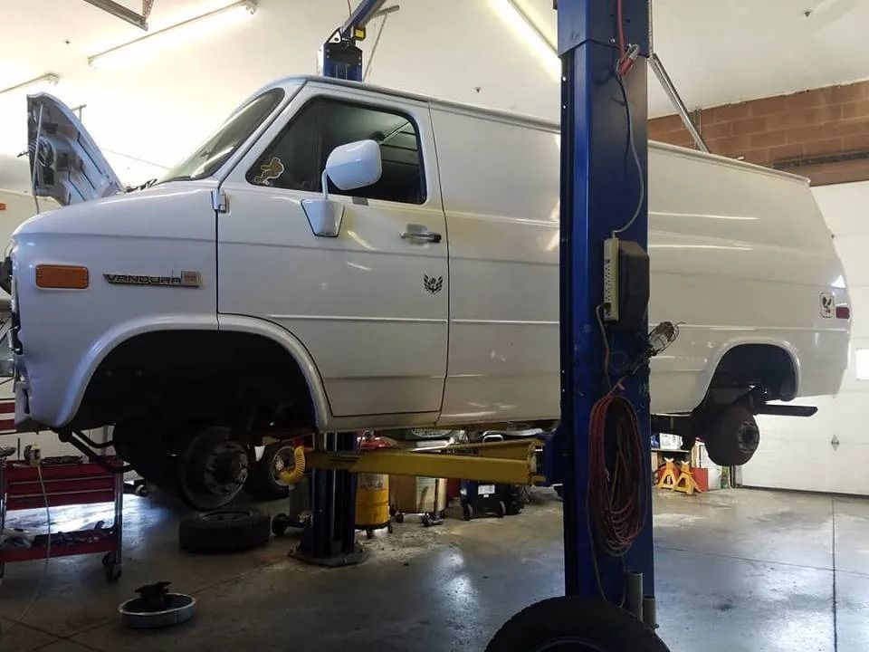 White van on a lift in a garage, wheels removed, hood open.