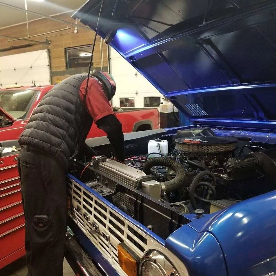 Mechanic working on the engine of a blue vintage car in a garage, wearing a black vest and red shirt.