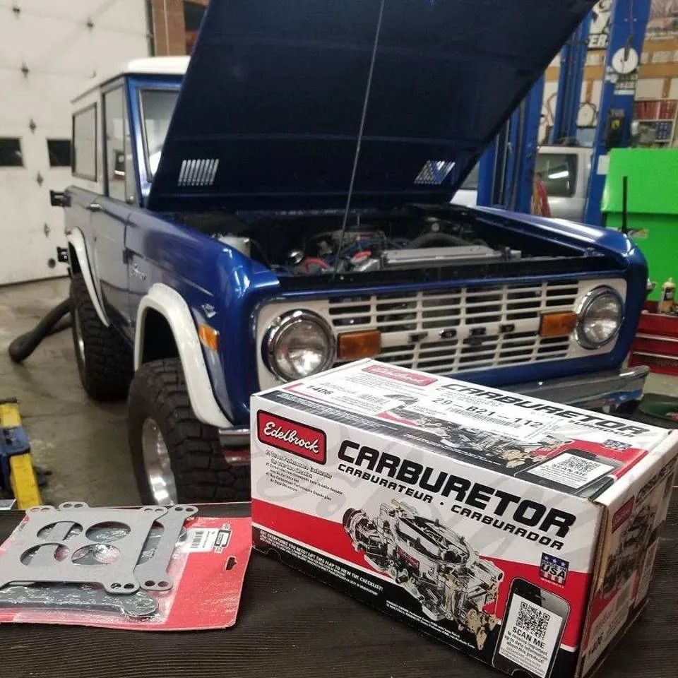 Blue classic Bronco with hood open, new carburetor box in front. Garage setting.