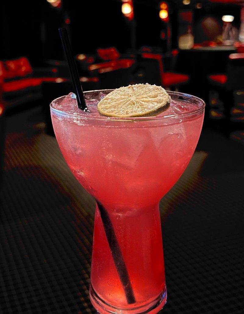 Pink cocktail in a stemmed glass, garnished with a citrus wheel, in a dimly lit bar setting.