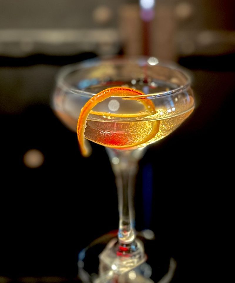 Cocktail in coupe glass with orange peel garnish, dark background.