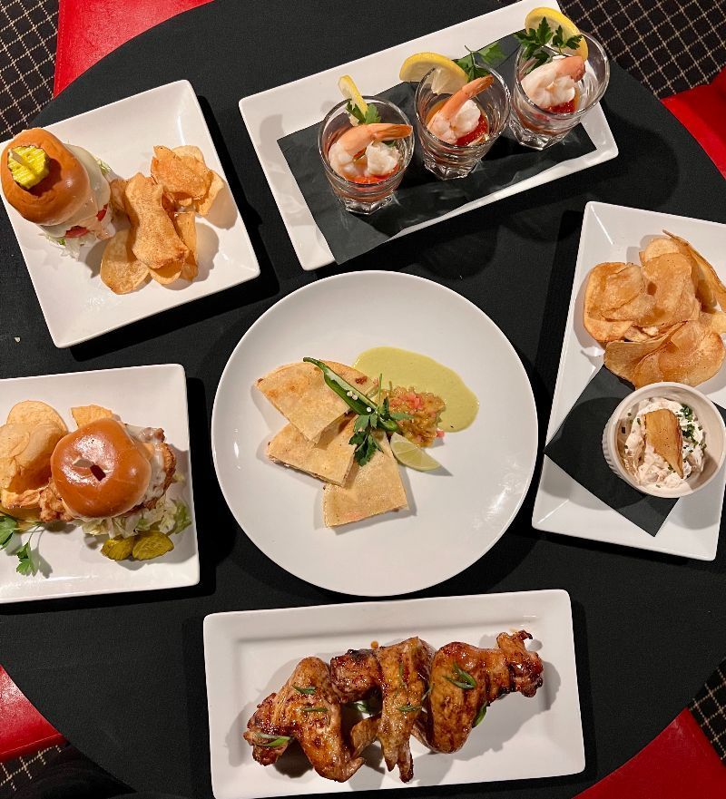 Assorted appetizers on a black table: shrimp cocktail, wings, sandwiches, chips, and a plate of squares, shot from above.