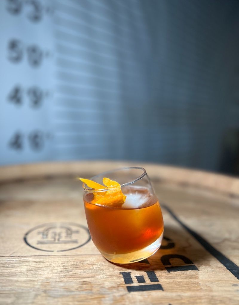 Cocktail with orange peel garnish on a wooden barrel, set against a blurred background with numbers.