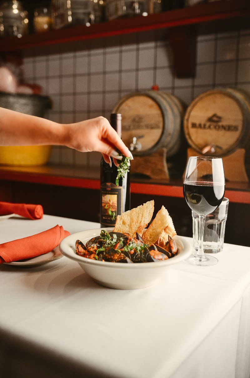 Hand sprinkling herbs over a bowl of food on a white tablecloth; wine and barrels in the background.