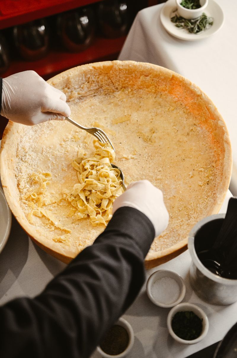 Chef tossing pasta in a large cheese wheel. White-gloved hands, several spices.