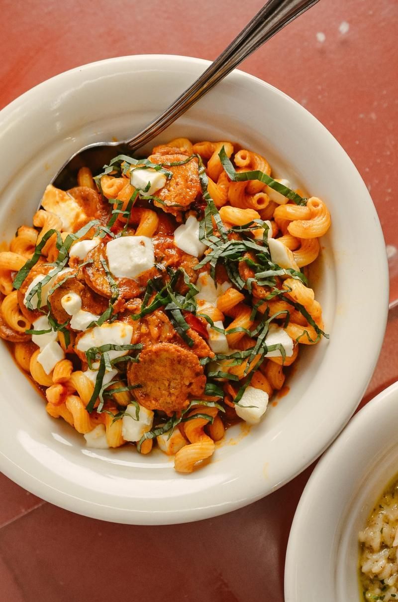 Pasta dish with sausage, mozzarella, and basil in a white bowl on a red table.
