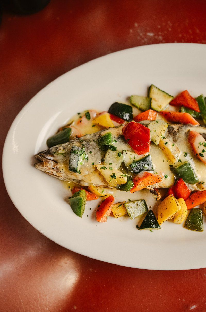 Baked fish with colorful vegetables on a white oval plate against a red background.