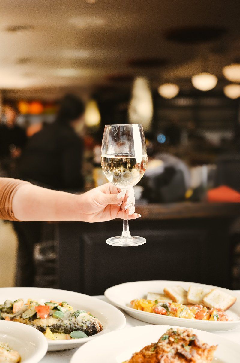 Person's hand holding a wine glass with food plates on a table in a restaurant.