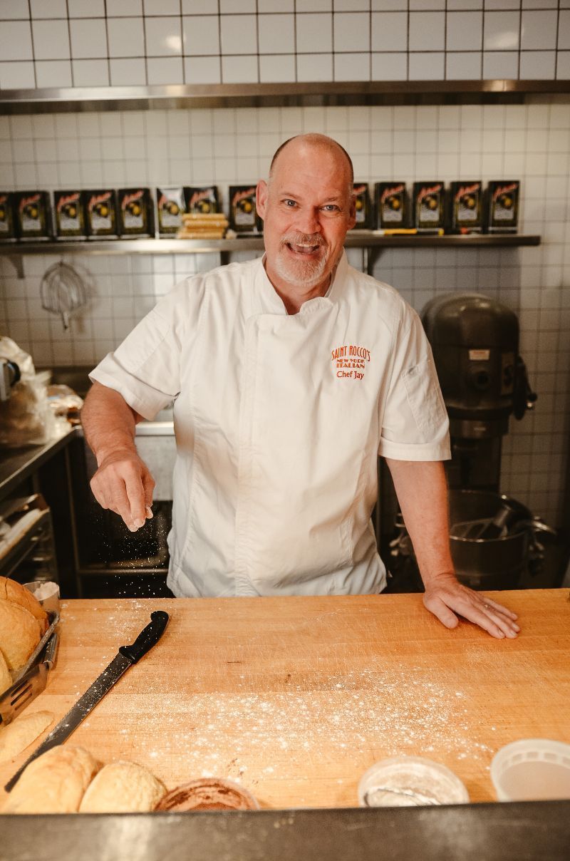 Baker in white coat, sprinkles flour, smiles at counter, in a bakery kitchen.