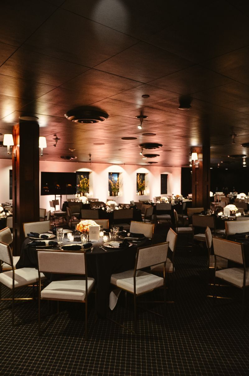 Dark event space with round tables set for dinner, white chairs, and muted lighting.