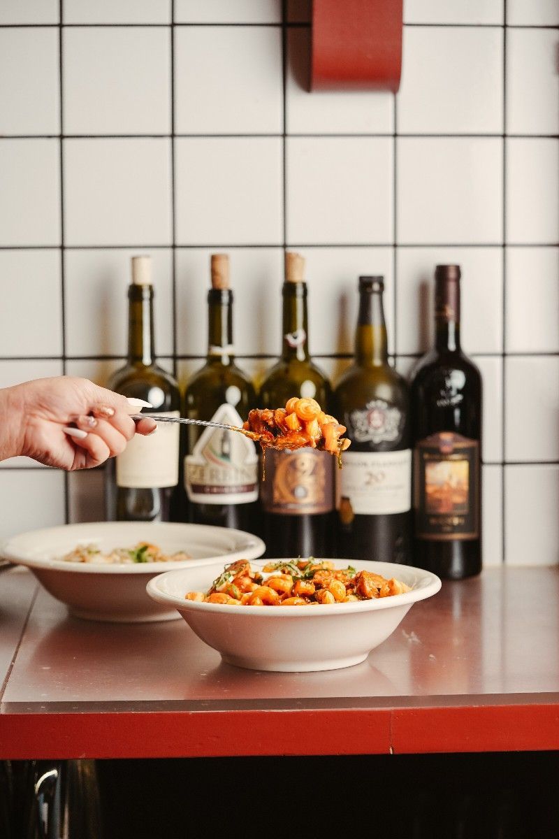 Pasta dishes, wine bottles, and a hand scooping food on a red counter in front of a tile wall.