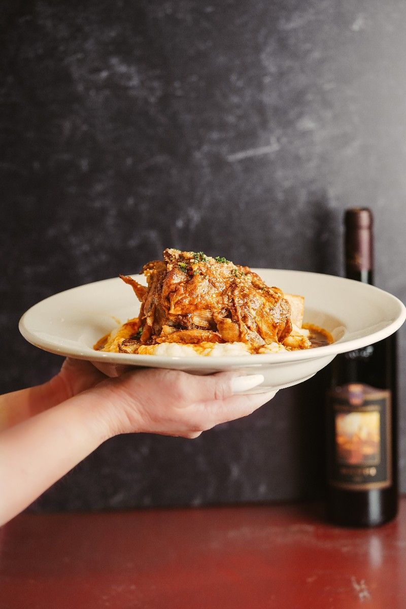 Hands holding a plate of chicken dish with pasta, next to a bottle of wine.