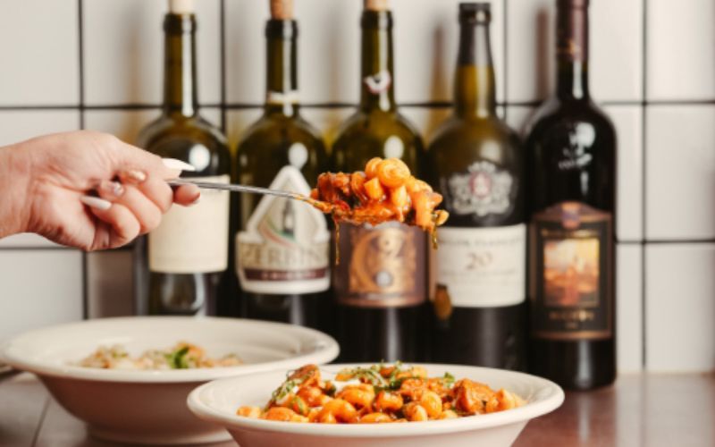 A person holds food on a fork above a bowl. Wine bottles are in the background.