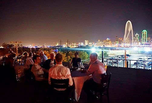 People dining outdoors at night with a city skyline, including a lit arch, in the background.