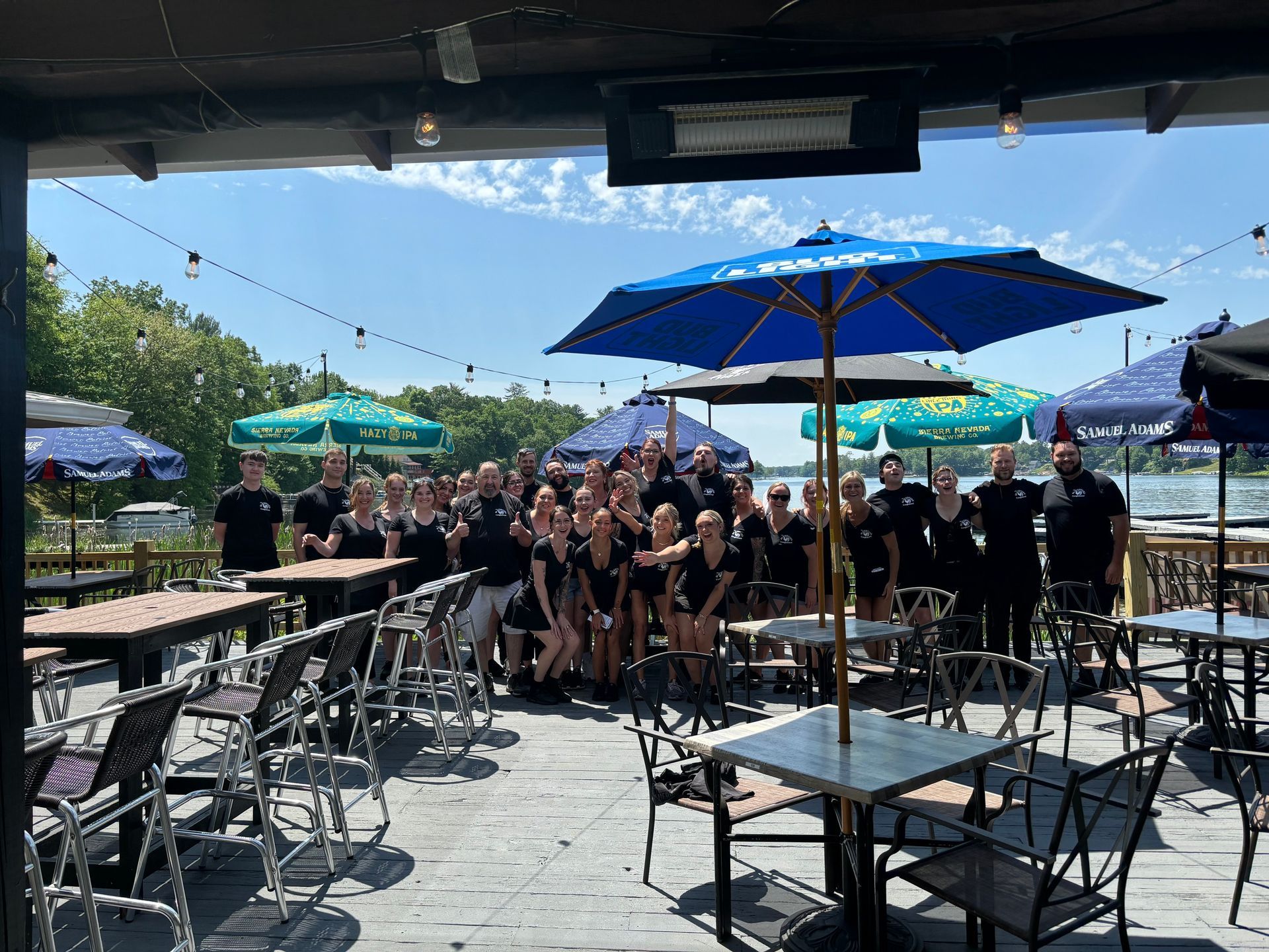 A group of people standing in front of tables and umbrellas on a patio.