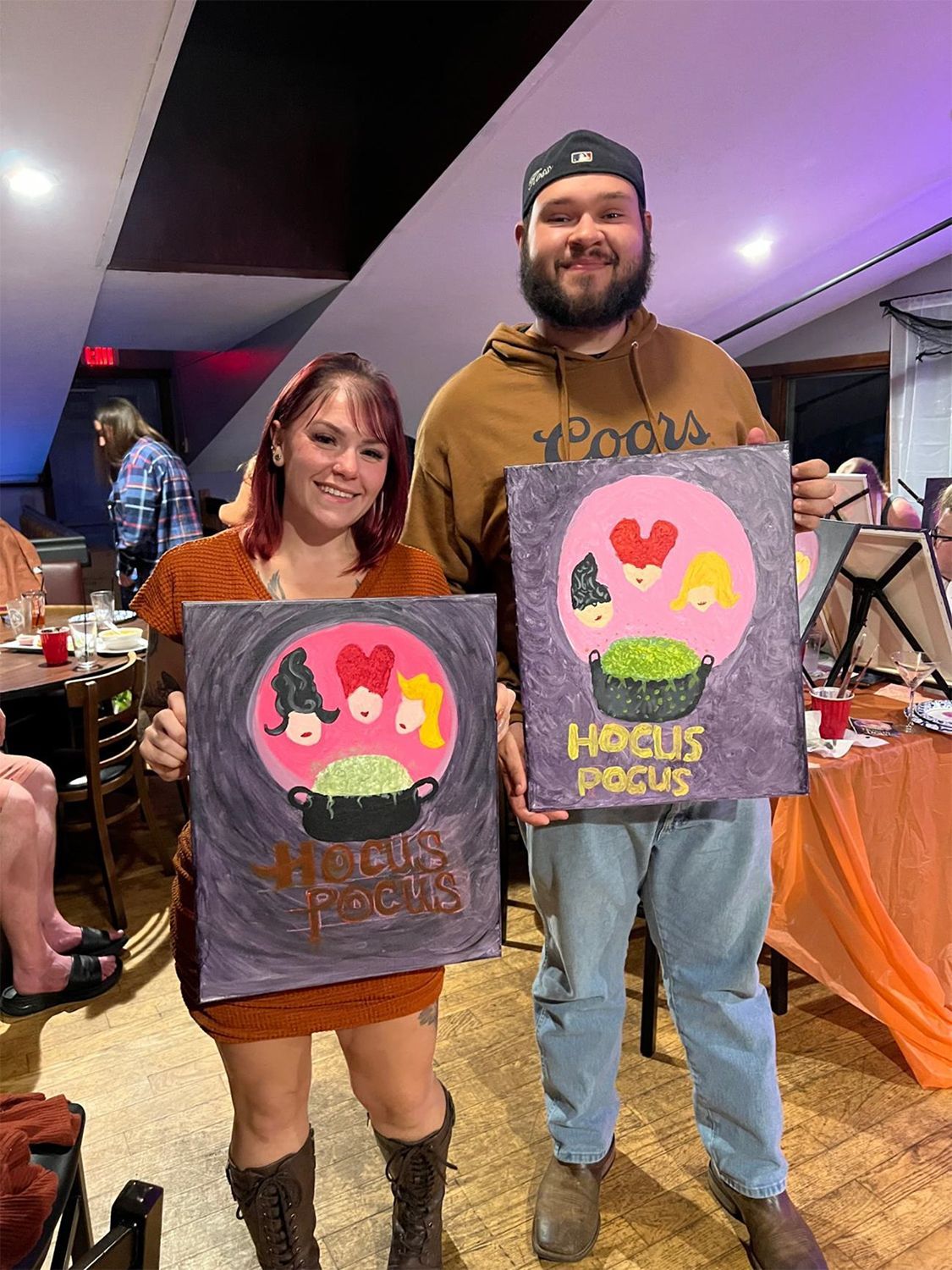 A man and a woman are standing next to each other holding two Halloween paintings.