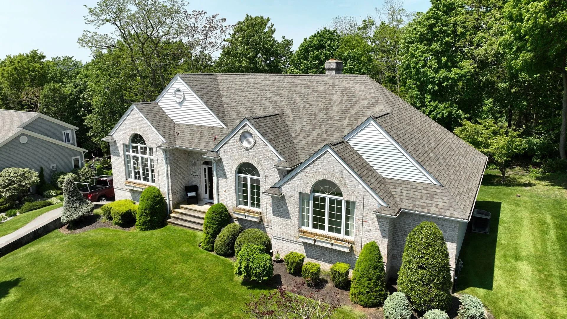 An aerial view of a gray stone house with a shingled roof, surrounded by green lawns and trees on a sunny day.