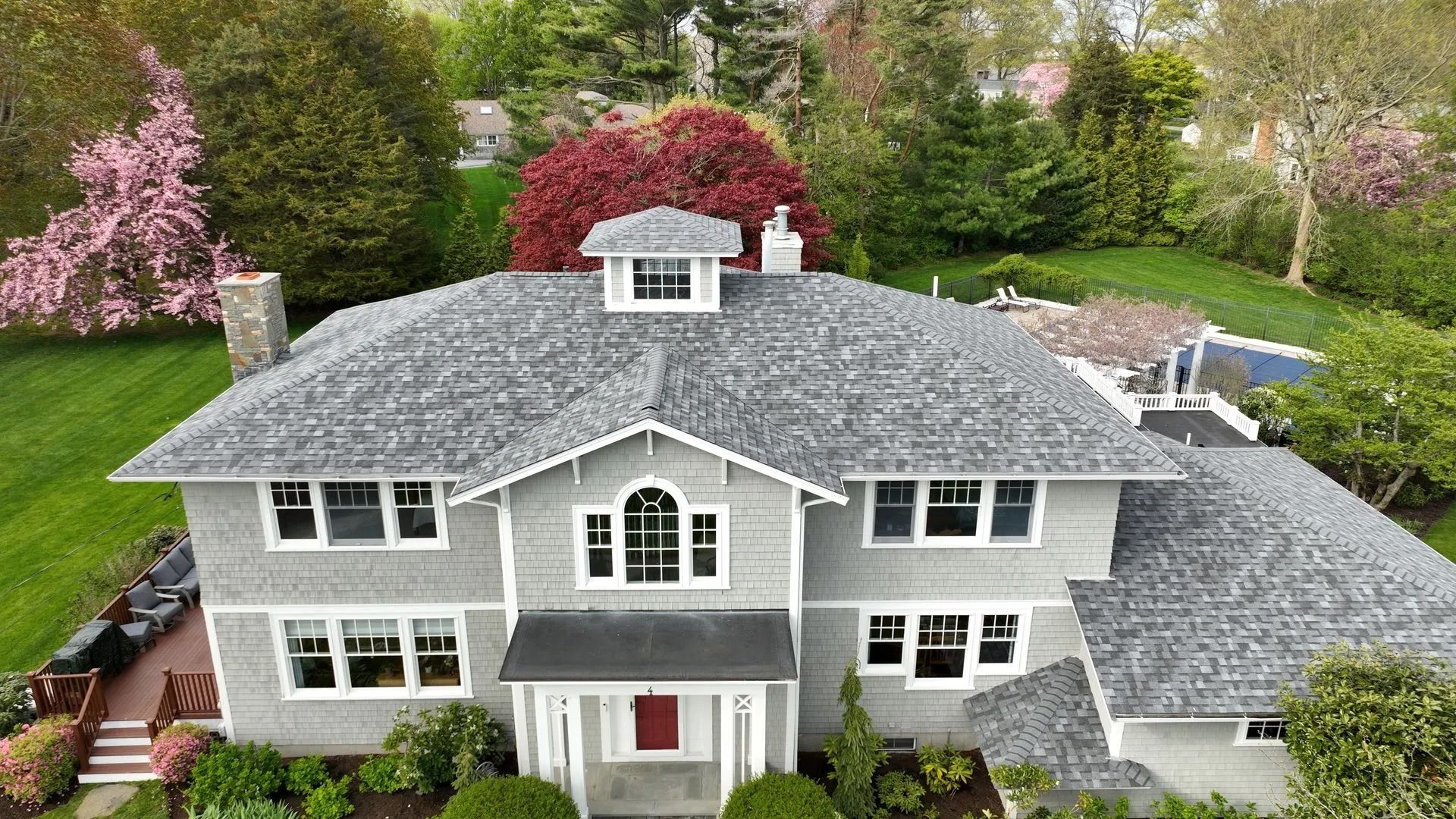 An aerial view of a gray, two-story house with a shingled roof, a central cupola, and a lush green landscape.